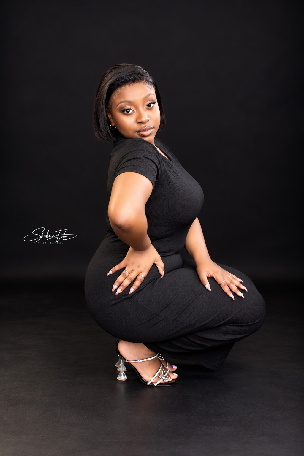Woman posing seated in a black dress against a dark studio backdrop